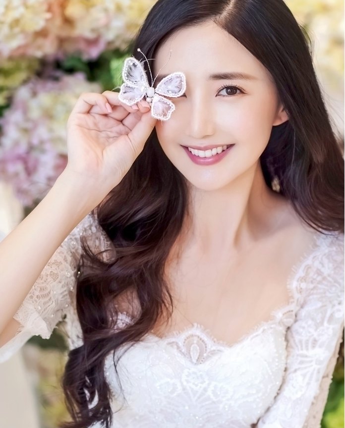 흰 레이스 드레스를 입은 여성이 흰 나비 장식을 눈 앞에 들고 미소 짓는 사진