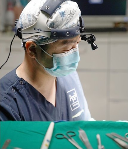 수술용 루페와 헤드라이트를 착용하고 마스크를 쓴 의료진이 수술에 집중하는 모습. 앞쪽 초록 소독포 위에 수술 기구가 놓여 있다.
