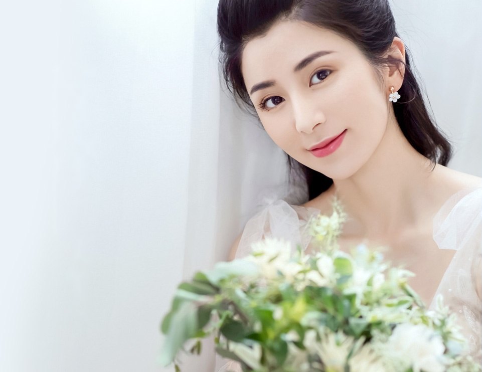 검은 긴 머리를 반묶음한 젊은 여성이 흰 드레스 차림으로 흰 꽃다발을 들고 옅게 미소 짓는 상반신 컷