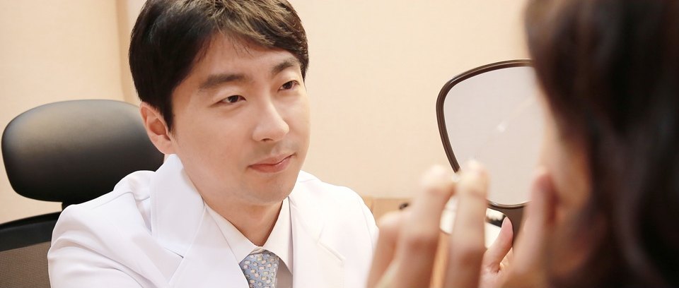흰 가운을 입은 의사가 손거울을 들고 환자와 쌍꺼풀 라인을 상담하는 모습