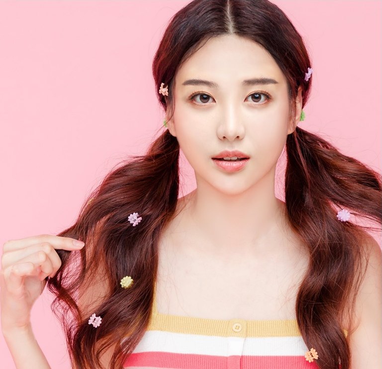 분홍 배경 스튜디오에서 양갈래로 묶은 긴 갈색 웨이브 머리에 꽃 모양 집게핀을 여러 개 꽂은 여성 모델이 정면을 보며 한 손으로 머리카락을 잡고 있는 모습